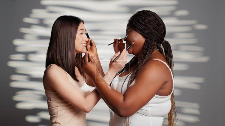 Photo for Interracial beauty models giving each other makeovers, using make up brushes and feeling cheerful in studio. Young friends enjoying fun skincare campaign, body positivity and acceptance. - Royalty Free Image