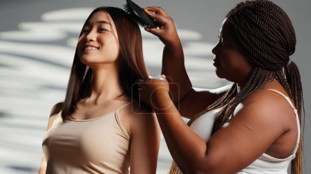 Téléchargez les photos : Diverses femmes riant et brossant les cheveux en studio, s'amusant comme des amis et montrant l'acceptation du corps. Jeunes filles diversifiées avec différents types de peau et de corps, campagne de soins de la peau. - en image libre de droit