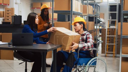 Mann im Rollstuhl arbeitet im Warenlager, Mitarbeiter mit chronischer Beeinträchtigung tragen Kisten, um Waren vor dem Versand zu kontrollieren. Kerl arbeitet in behindertengerechtem Raum.