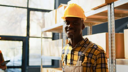 Photo for African american man posing next to boxes on racks, working in warehouse with retail products for shipment. Male storage room worker drinking coffee before checking merchandise. Handheld shot. - Royalty Free Image
