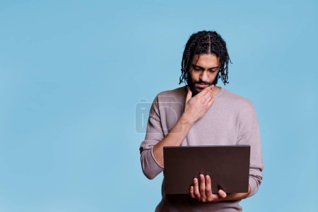 Photo for Young thoughtful arab man holding laptop while analyzing software code and rubbing chin. Pensive person thinking in doubt while reading business report on portable computer - Royalty Free Image