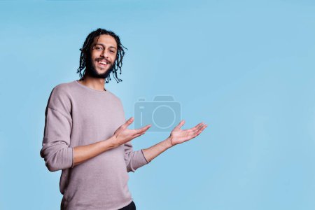 Téléchargez les photos : Homme arabe souriant pointant avec les mains sur le côté tout en annonçant le produit et en regardant la caméra. Personne joyeuse avec une expression faciale positive montrant à gauche avec des bras portrait - en image libre de droit
