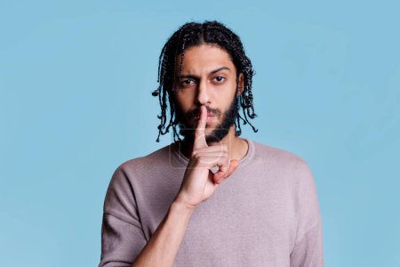 Photo for Arab man making quiet gesture and holding forefinger on lips portrait. Young person with serious facial expression doing tsss sign and looking at camera on blue background - Royalty Free Image