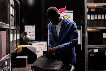 Téléchargez les photos : Un comptable afro-américain met son sac à dos, quitte le travail tard dans la nuit. Homme d'affaires travaillant des heures supplémentaires au dossier de la bureaucratie, l'analyse des documents administratifs dans la salle d'entreposage. - en image libre de droit