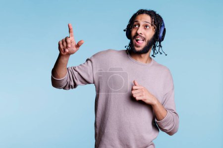 Foto de Hombre árabe feliz en auriculares inalámbricos bailando, moviendo las manos y señalando con los dedos. Joven bailarina alegre divirtiéndose y disfrutando de la música en auriculares sobre fondo azul - Imagen libre de derechos