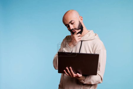 Foto de Pensativo árabe hombre frotando la barbilla en duda mientras trabaja en el ordenador portátil. Joven persona pensativa con el pensamiento de expresión perplejo mientras sostiene el ordenador portátil y el análisis de datos de negocios - Imagen libre de derechos