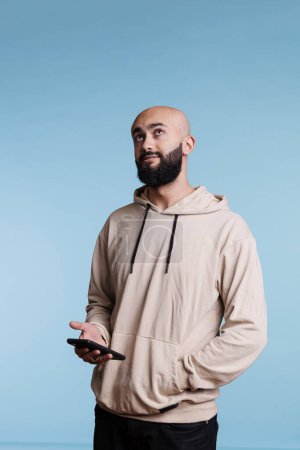 Foto de Hombre árabe usando un teléfono inteligente, pensando mientras escribe un mensaje en línea y mirando hacia arriba. Joven con expresión facial pensativa sosteniendo el teléfono móvil mientras está de pie con la mano en el bolsillo con capucha - Imagen libre de derechos