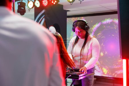 Photo for Woman dj in headphones mixing music on stage during electronic music concert in nightclub. African american musician in earphones performing on at discotheque party in club - Royalty Free Image