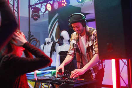 Dj in headphones using jog wheel and knobs while mixing sound with controller on nightclub stage. Young man musician playing electronic music during disco party in dark club