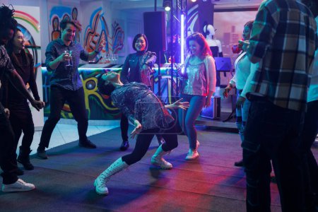 Friends watching woman dancing in nightclub, laughing while attending disco party. Diverse happy people crowd enjoying night discotheque on dancefloor with spotlights in club