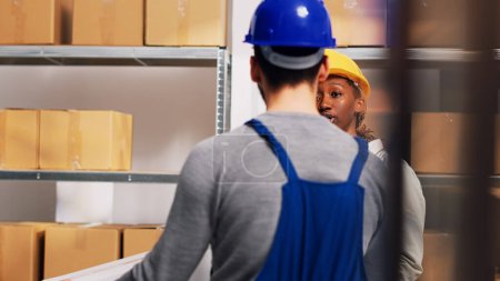 Photo for African american supervisor looking at boxes of goods in depot, checking stock logistics in warehouse shelves. Man and woman working with merchandise placed in boxes on depot racks. Handheld shot. - Royalty Free Image