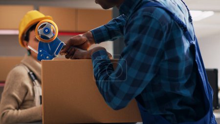 Photo for African american man putting adhesive tape on boxes, preparing order with warehouse merchandise in storage room. Male employee sealing off packages with products, delivery service. - Royalty Free Image