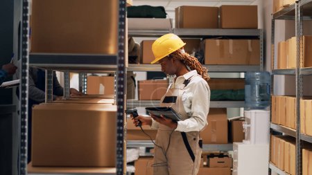 Photo for African american woman scanning barcodes on boxes, using digital tablet and scanner to ship manufacturing goods. Female supervisor working with warehouse products on depot shelves. Tripod shot. - Royalty Free Image