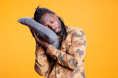 Téléchargez les photos : Homme afro-américain mettre la tête sur un oreiller confortable pour dormir et faire la sieste. Personne épuisée essayant de se reposer après avoir passé l'heure du coucher, dormant en studio sur fond jaune. - en image libre de droit