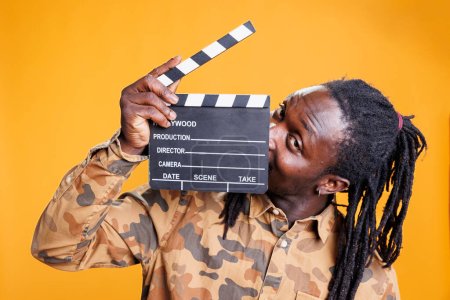 Photo for Portrait of smiling african american man holding movie production blackboard standing in studio over yellow background. Cheerful person giving audition for entertainment video. Cinema concept - Royalty Free Image