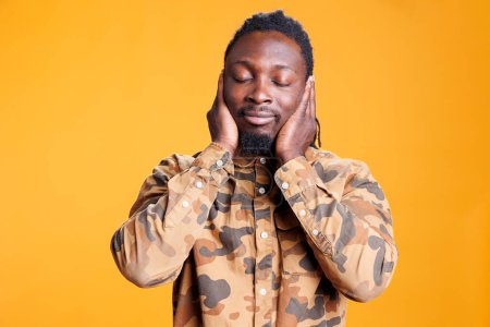 Man with close eyes covering ears with palms, doing three wise monkeys gesture in front of camera over yellow background. African american person not listening to noise and not speaking in studio
