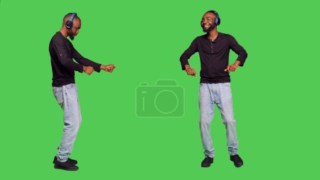 Téléchargez les photos : Modèle masculin avec écouteurs dansant à la caméra, écoutant de la musique sur casque audio et s'amusant. Jeune faisant des mouvements de danse alors qu'il aime les chansons positives, debout sur l'écran vert. - en image libre de droit