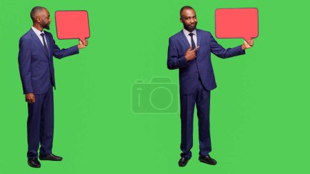 Photo for Startup employee holding blank speech bubble, using empty isolated copyspace for advertisement in studio. Businessman in formal suit showing red billboard for ad on full body greenscreen. - Royalty Free Image