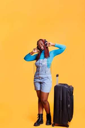 Photo for Woman with trolley bag and headphones listening to music and enjoying holiday trip, going on vacation with big suitcase. Young adult having fun with audio headset in studio, travelling. - Royalty Free Image