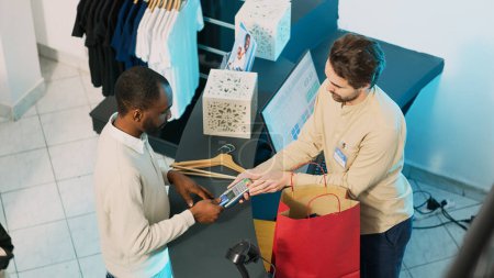 Photo for Male shopper buying merchandise at cash register, using credit card to make payment on pos terminal. Fashion boutique worker giving shopping bags to client, commercial activity. Handheld shot. - Royalty Free Image