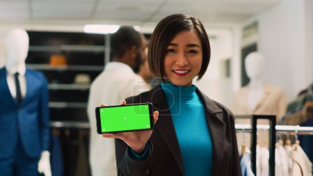 Foto de Mujer asiática sosteniendo pantalla verde aislada, mostrando la plantilla de croma clave en la tienda de ropa. Empleada de la tienda usando espacio en blanco aislado en la pantalla del teléfono móvil. - Imagen libre de derechos