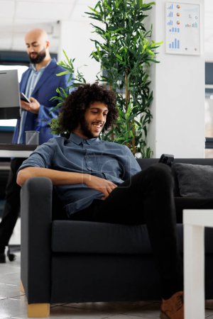 Téléchargez les photos : Travailleur de start-up souriant regardant la vidéo sur smartphone pendant la pause de travail dans le bureau d'affaires. Entrepreneur arabe joyeux utilisant le téléphone mobile et se détendre dans l'espace de coworking - en image libre de droit