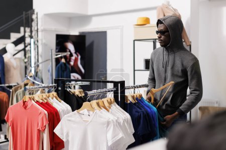 Photo for African american man thief taking a risk, looking at trendy clothes before stealing it in modern boutique. Robber wearing sunglasses and hood not be recognized in shopping centre. Burglary concept - Royalty Free Image