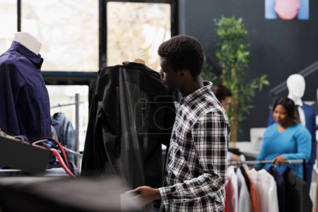 Foto de Cliente afroamericano que busca comprar camisa formal, comprobando la tela de mercancía en la tienda de ropa. Elegante hombre de compras ropa de moda en la boutique moderna. Concepto de moda - Imagen libre de derechos