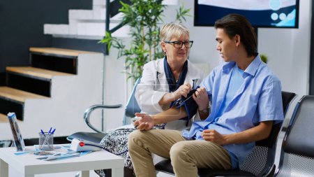 Photo for Senior traumatologist using tonometer to check patient high or low pulse pressure during medical appointment in hospital waiting area. Elderlt doctor measuring asian man hypertension and hypotension - Royalty Free Image