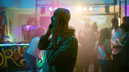Person answering phone call at nightclub, trying to have remote conversation at disco party. Man talking on smartphone, enjoying night out at event with funky music. Handheld shot.