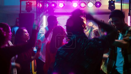 Foto de Hombres y mujeres jóvenes saltando en el evento, sintiéndose felices festejando con música funky. Multitud de gente feliz bailando y sintiéndose bien en la reunión social, disfrutando de la vida nocturna. Disparo de mano. - Imagen libre de derechos