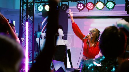 Foto de Mujer joven trabajando como DJ en la fiesta y mezclando música en la estación estéreo, divirtiéndose en el escenario con focos. Funky persona bailando y saltando con la gente en el club nocturno. Disparo de mano. - Imagen libre de derechos