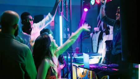 Foto de Grupo multiétnico de adultos festejando y bailando en la música del club, divirtiéndose en la discoteca con DJ en el escenario. Hombres y mujeres saltando alrededor de la pista de baile y sintiéndose felices en el evento. Disparo de mano. - Imagen libre de derechos
