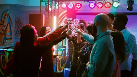 Photo for Smiling people clinking glasses filled with alcohol on dance floor, saying cheers under colorful spotlights. Young men and women enjoying cool funky party, entertainment. Handheld shot. - Royalty Free Image