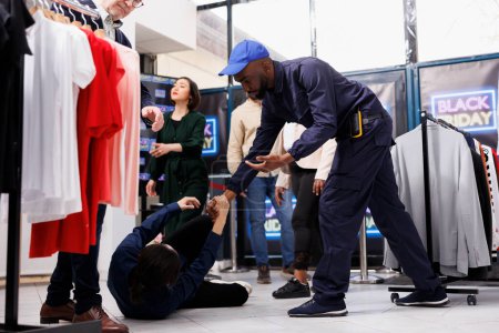 Freundlicher afroamerikanischer Typ, Sicherheitsbeamter eines Bekleidungsgeschäfts, der Einkäufern während des Black-Friday-Wahnsinns hilft. Junger Mann fällt beim Einkaufen in Einkaufszentrum während Saisonverkauf auf den Boden