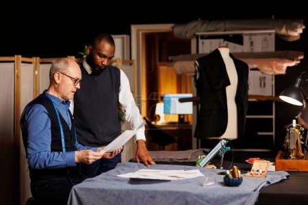 Photo for Skilled tailors comparing different fashion sketches attire combinations in atelier workspace. Dressmaker and apprentice working on clothing alterations, preparing for upcoming collection release - Royalty Free Image