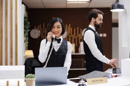 Photo for Asian employee at hotel reception desk, working on booking rooms and filling in reservations record. Female receptionist answering landline phone call in resort lobby, accommodation. - Royalty Free Image