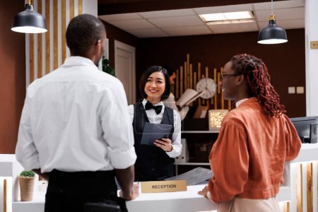 Photo for Receptionist registering guests in lobby, filling in hotel record files at reception counter desk. Asian employee helping people with registration to give room key card, welcome tourists. - Royalty Free Image