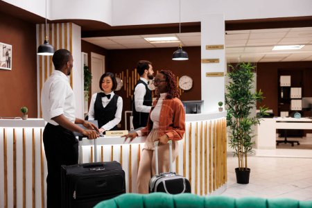 Photo for Couple arriving at hotel reception with luggage, talking to receptionist at front desk about accommodation. Man and woman waiting to do check in with room reservation, registration. - Royalty Free Image