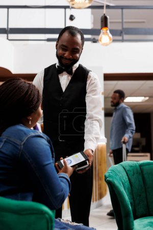 Téléchargez les photos : Serveur souriant afro-américain donnant pos machine à invité de l'hôtel féminin assis dans le hall. Un smartphone touristique payant pour commander en attendant l'enregistrement dans le salon - en image libre de droit