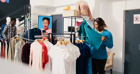 Foto de Empleada asiática ayudando a la mujer a comprar clohtes elegantes, discutiendo el material de la camisa en la tienda de ropa. cliente afroamericano de compras de mercancías de moda, mirando a la nueva colección de moda - Imagen libre de derechos