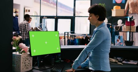 Foto de Manager mirando a la computadora con pantalla verde croma clave simulan pantalla, de pie en el mostrador en la boutique moderna. Empleado esperando clientes en la tienda de ropa. Concepto de moda - Imagen libre de derechos