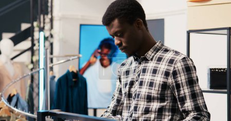 Téléchargez les photos : Acheteur afro-américain regardant une nouvelle collection de mode, analysant le tissu de marchandises dans une boutique moderne. Achat de vêtements à la mode dans un magasin de vêtements - en image libre de droit
