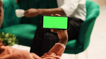 Téléchargez les photos : Femme afro-américaine tenant chroma écran vert mock up smartphone en mode paysage assis sur un canapé confortable. Touristes en attente dans l'élégant salon de l'hôtel à enregistrer - en image libre de droit