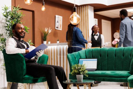 Photo for Friendly African American guy hotel porter sitting in lobby with clipboard and laptop. Positive cheerful black man bellhop smiling to guests while making entry in daily luggage movement register - Royalty Free Image