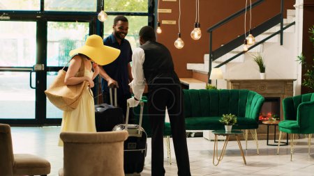 Photo for Hotel bellhop providing luxury service to guests entering reception lobby, concierge carrying baggage and trolley bags to room. Young couple arriving at tropical resort front desk. - Royalty Free Image