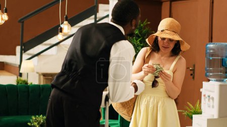 Photo for Couple giving cash tip to bellboy, receiving luxury concierge service with employee carrying baggage and trolley bags. Hotel guests paying money to male worker in lobby. Handheld shot. - Royalty Free Image