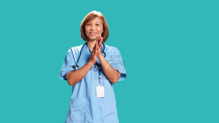 Photo for Approving smiling nurse happily clapping hands, satisfied with accomplishments. Healthcare specialist cheering, doing praising gesture while at work, isolated over blue studio background - Royalty Free Image