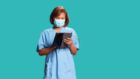 Téléchargez les photos : Infirmière asiatique vivante regardant les données des patients sur tablette numérique, confondue avec les résultats des examens médicaux. Travailleur clinique professionnel portant un masque protecteur, isolé sur fond bleu studio - en image libre de droit