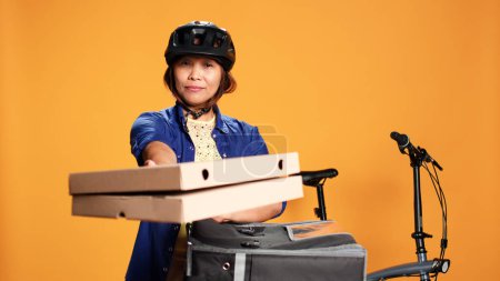 Téléchargez les photos : Courier femme isolée sur fond de studio orange apportant de la pizza au client. BIPOC cycliste livrer l'ordre au client, prendre des boîtes de restauration rapide hors du sac à dos thermique - en image libre de droit